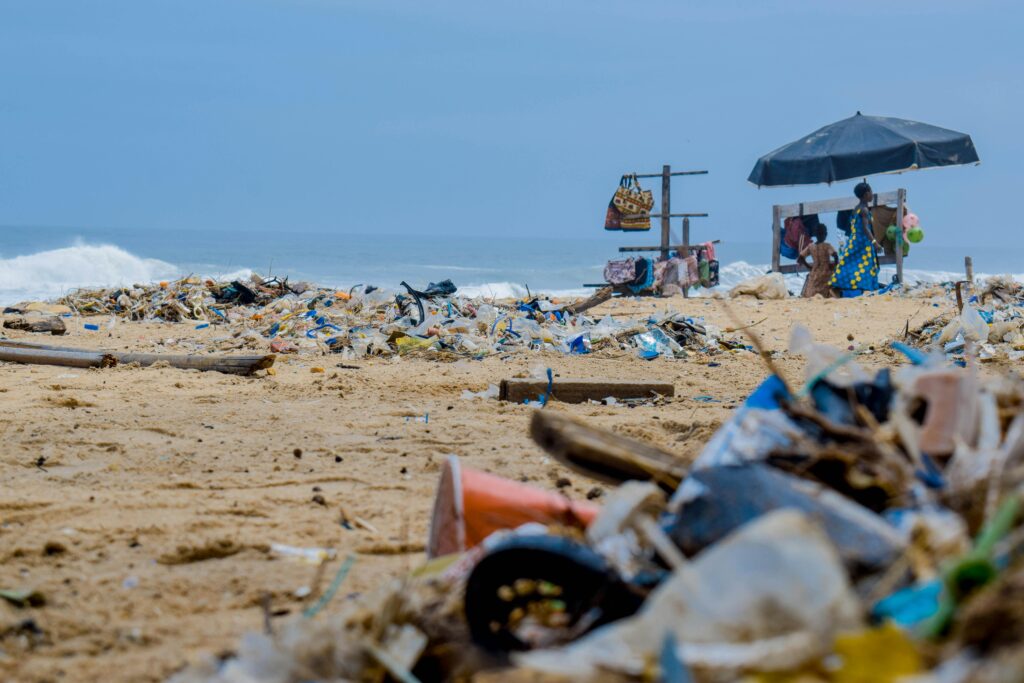 Sandy beach cluttered with trash and ocean debris, with vendors under an umbrella nearby.